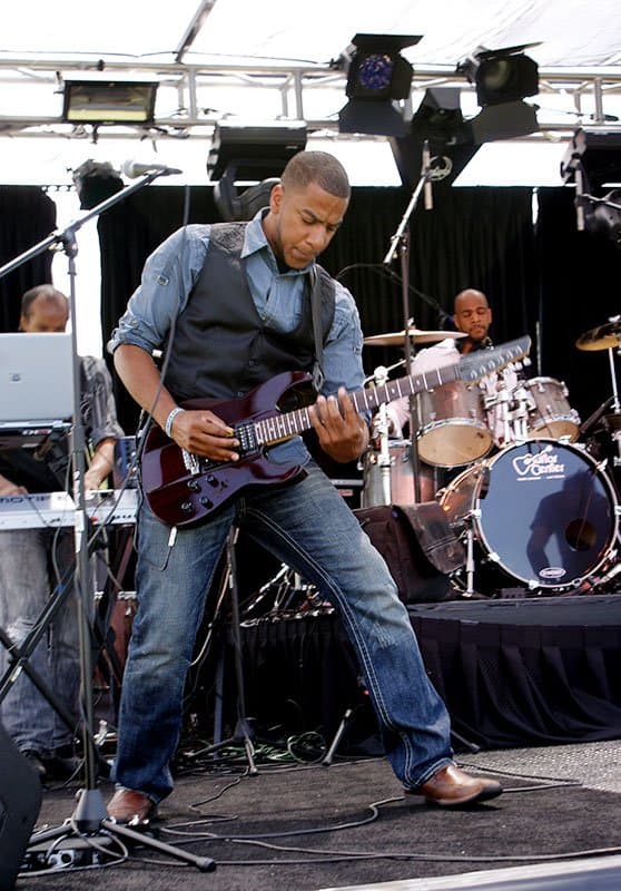 Guitarist performing on an outdoor stage with a drummer and keyboardist behind him