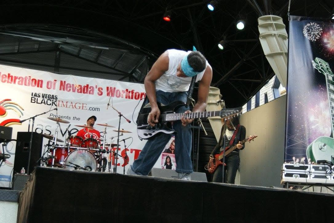 Singer in a white shirt and leopard hat performing on an outdoor stage with a keyboardist in the background
